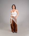 Woman wearing a white strapless top and brown wide-leg pants with a beige netted wrap on a gray background