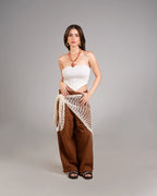 Woman wearing a white strapless top and brown wide-leg pants with a beige netted wrap on a gray background