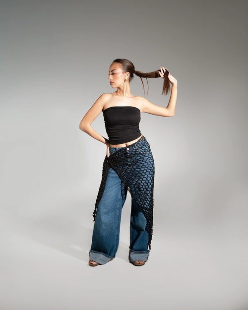 Woman wearing a black strapless top and blue wide-leg pants with a black netted accessory on a gray background