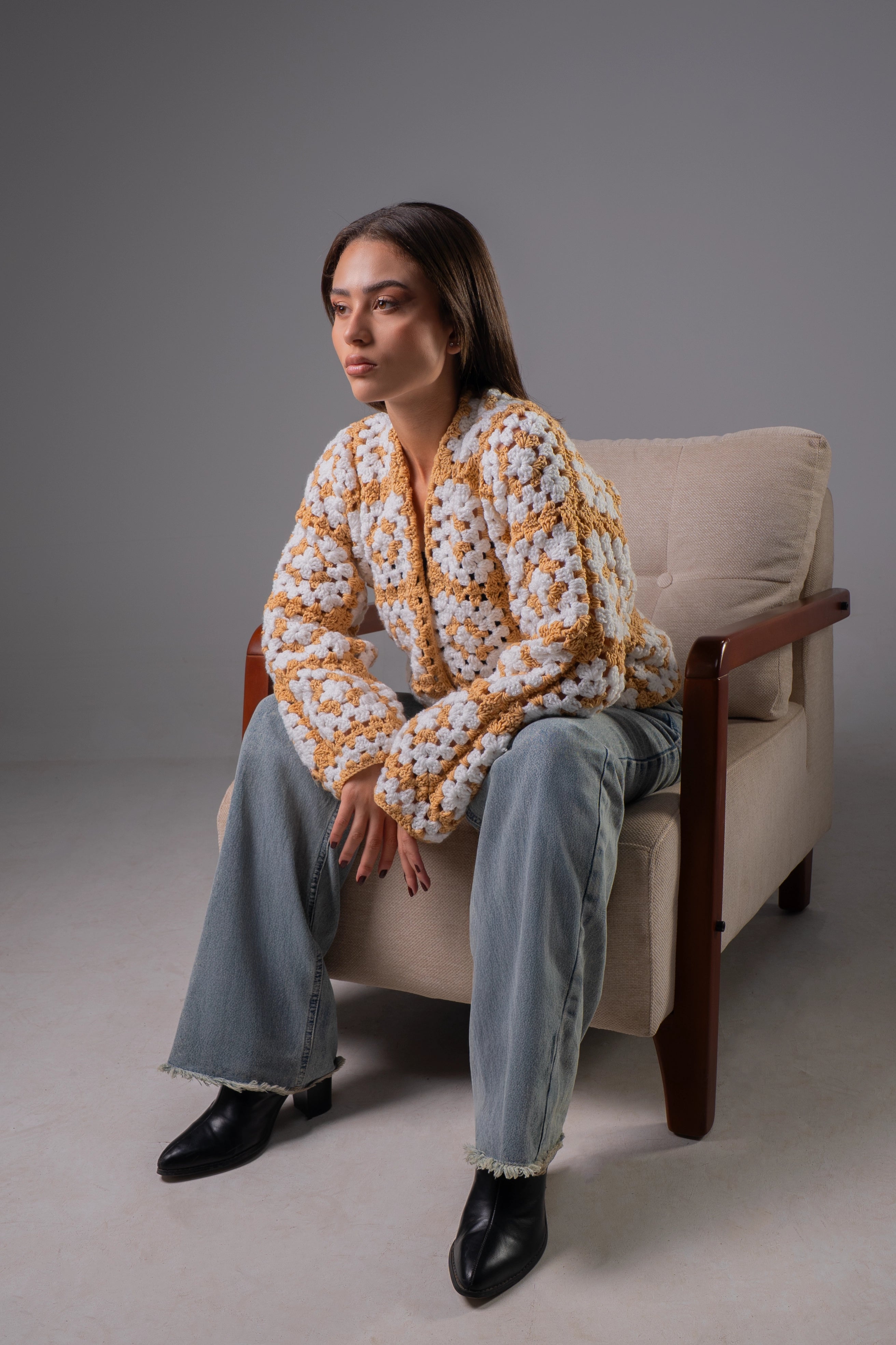 Woman sitting on a beige armchair wearing a patterned sweater and jeans against a gray background
