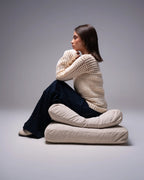 Woman sitting on a stack of cushions wearing a large beige knitted sweater against a gray background