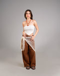 Woman wearing a white strapless top and brown wide-leg pants with a beige netted wrap on a gray background