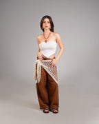 Woman wearing a white strapless top and brown wide-leg pants with a beige netted wrap on a gray background