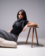 Woman sitting on a stool wearing a black top and gray scarf against a plain background