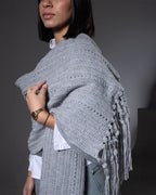 Person wearing a gray crocheted shawl with fringe on a dark background