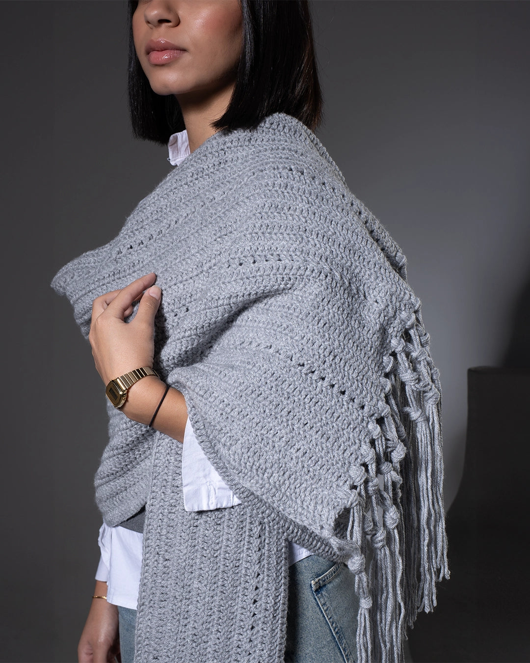 Person wearing a gray crocheted shawl with fringe on a dark background