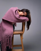 Woman wearing a purple knitted poncho sitting on a wooden stool against a gray background