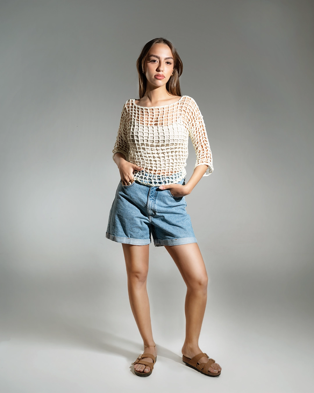 Woman wearing a beige crochet top and blue shorts on a gray background