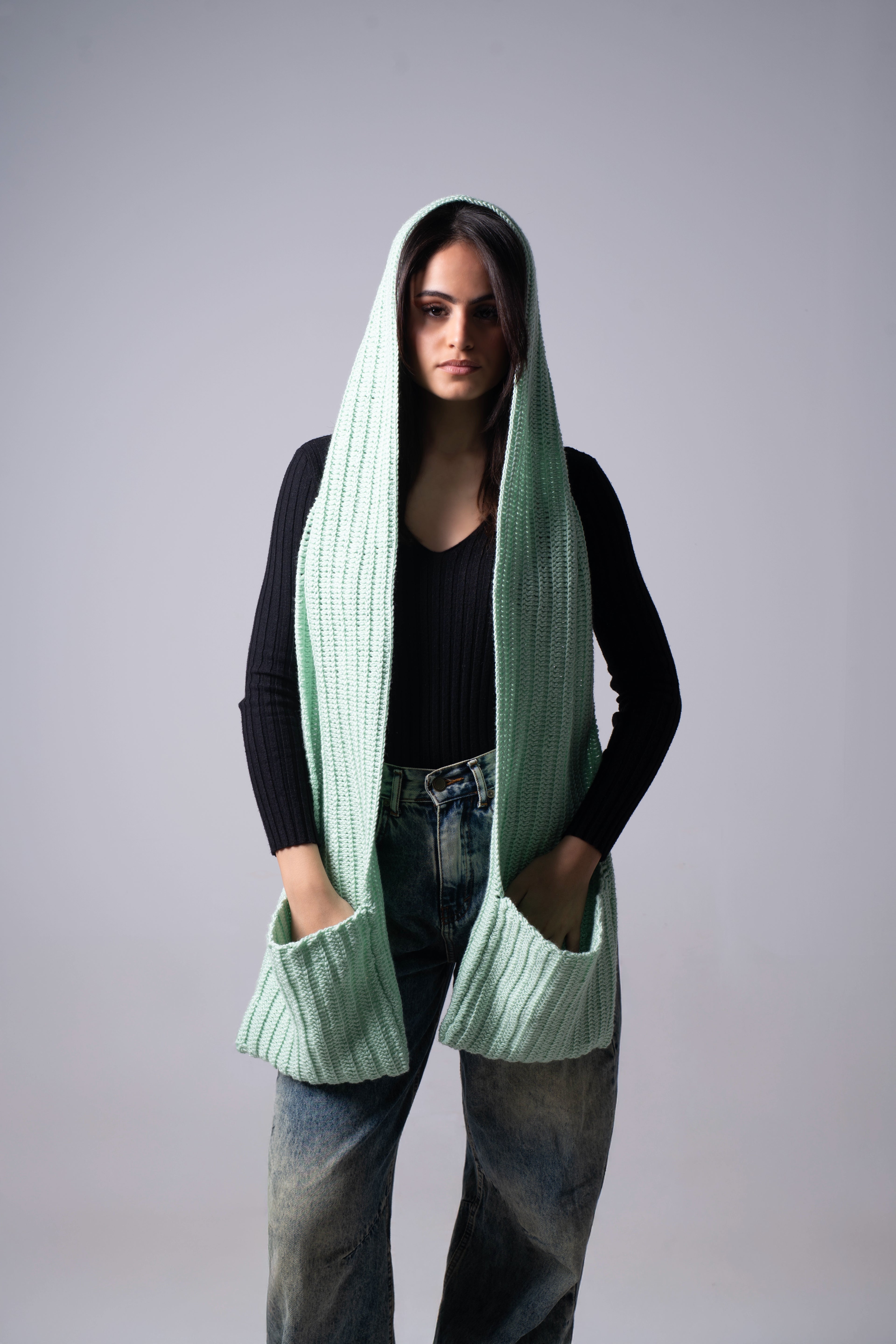 Person wearing a long green knitted scarf with a hood on a gray background