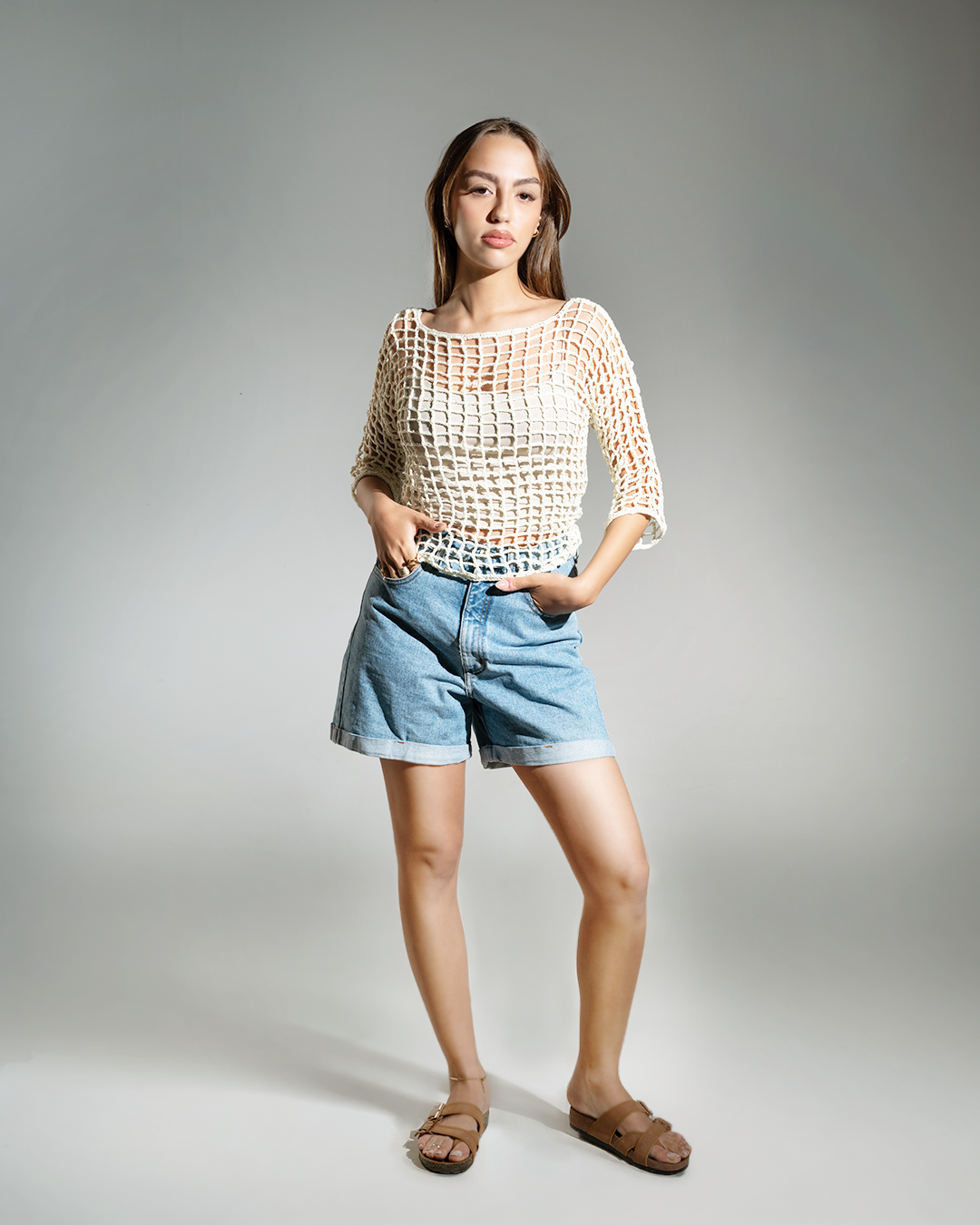 Woman wearing a beige crochet top and blue shorts on a gray background