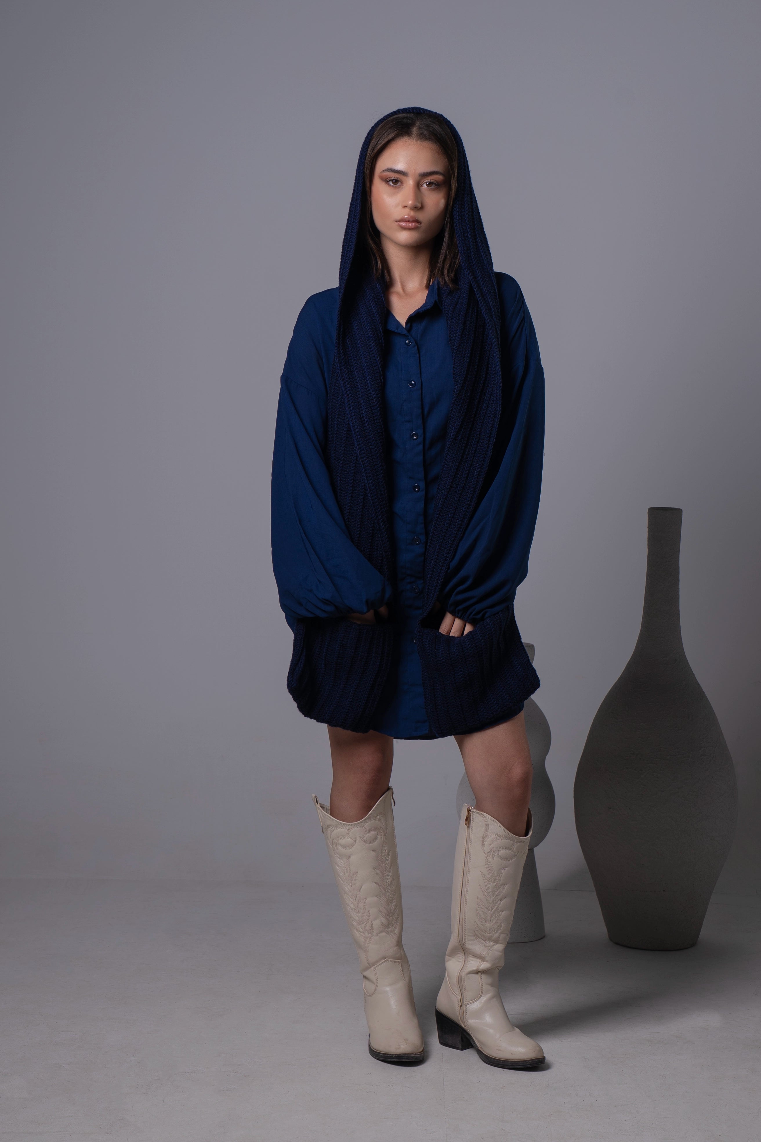 Woman wearing a blue dress with a hood and white boots standing against a gray background